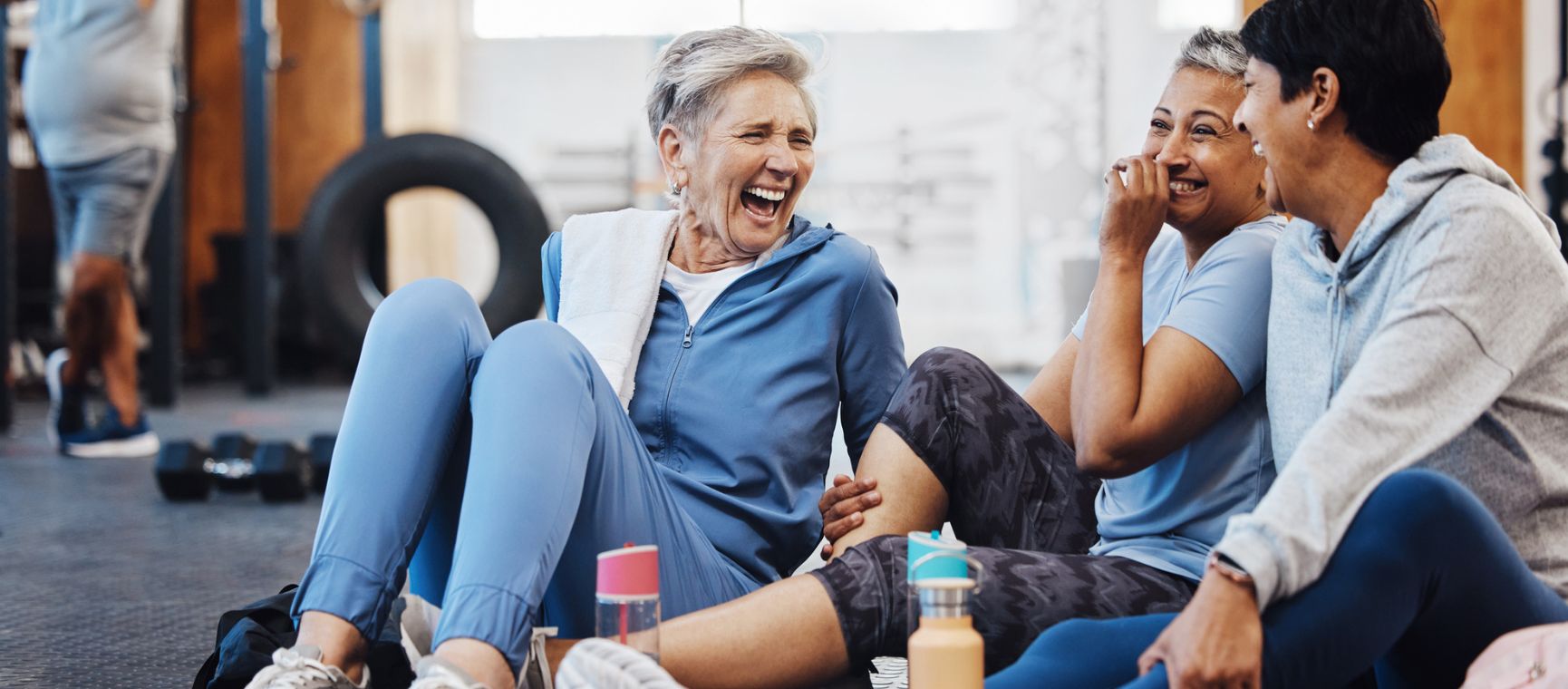 Three woman laughing and chatting sat on the floor of a gym wearing gym kit