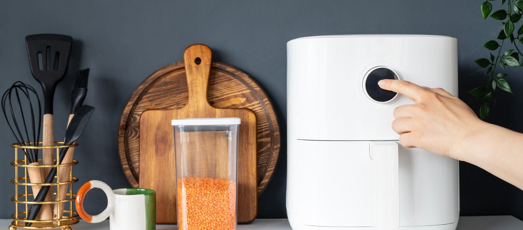 An air fryer on a counter next to kitchen implements with someone pressing the start button