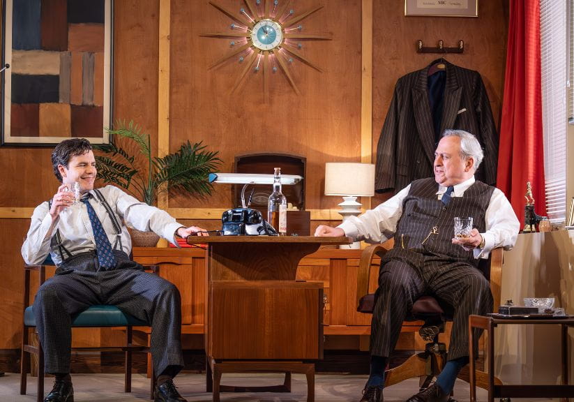 Two actors on stage around the desk in an office