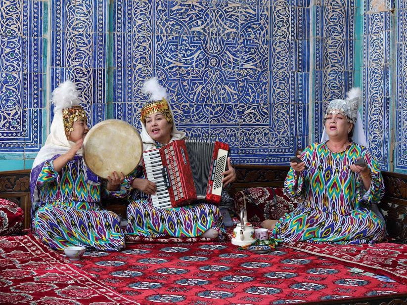 3 women sat on a decorative rug playing musical instruments against a decorative wall