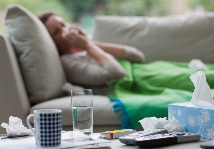 A woman ill with flu on her sofa with a box of tissues on the table in front of her
