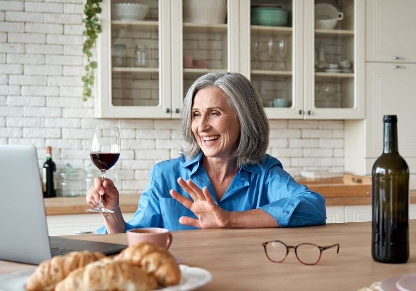 A woman holding a glass of wine and smiling and waving at someone on a laptop