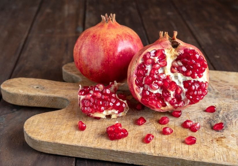 Two pomegranates with one cut in half showing its seeds and flesh on a wooden chopping board