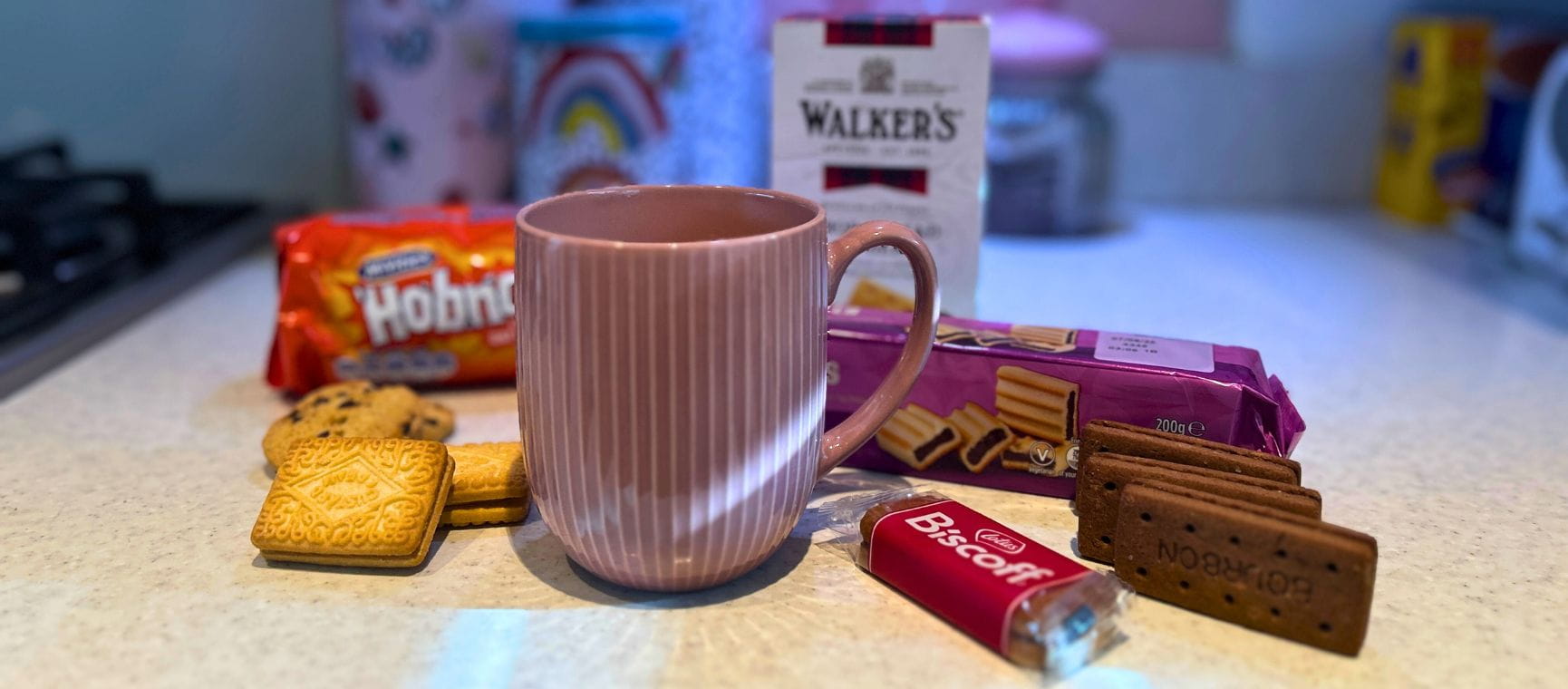 A line up of various biscuits in their packets and loose next to a cup of tea on a kitchen worktop to illustrate the best biscuits