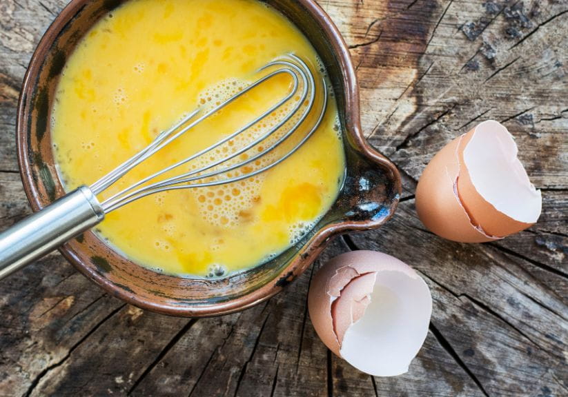A flat lay of some whisked egg in a bowl, with a whisk, alongside some broken eggs on a table