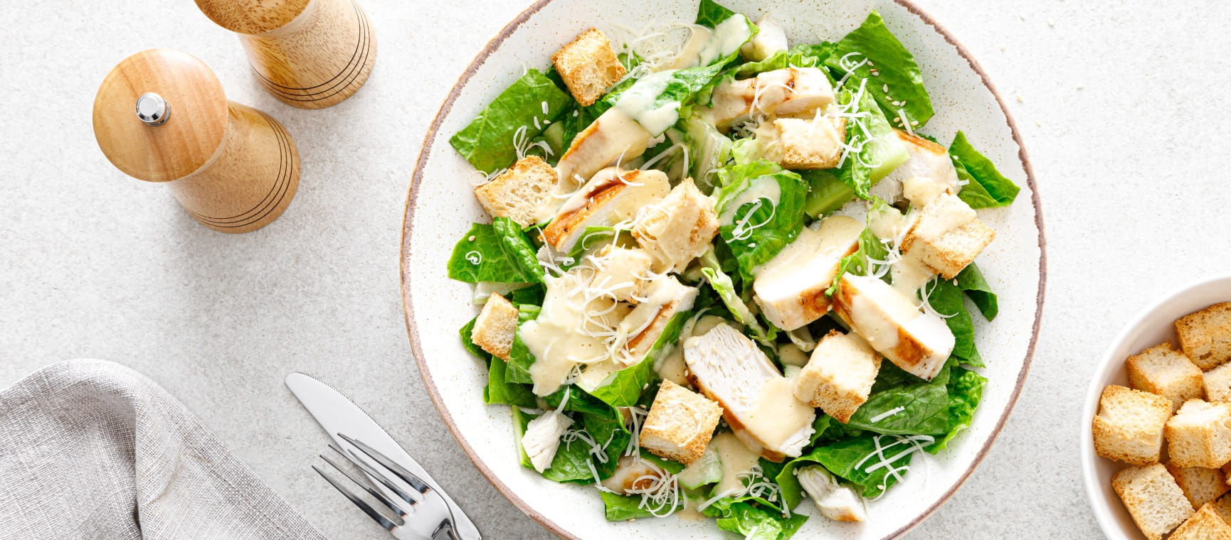 A bowl of chicken Caesar salad with a serving of croutons and condiments on the side