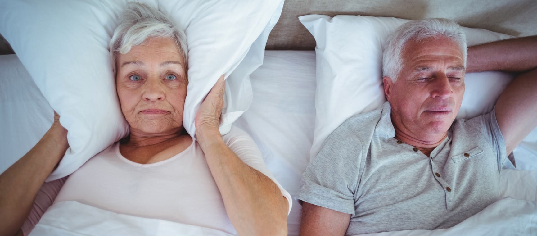 An older woman covering her ears with a pillow in bed while her husband snores next to her