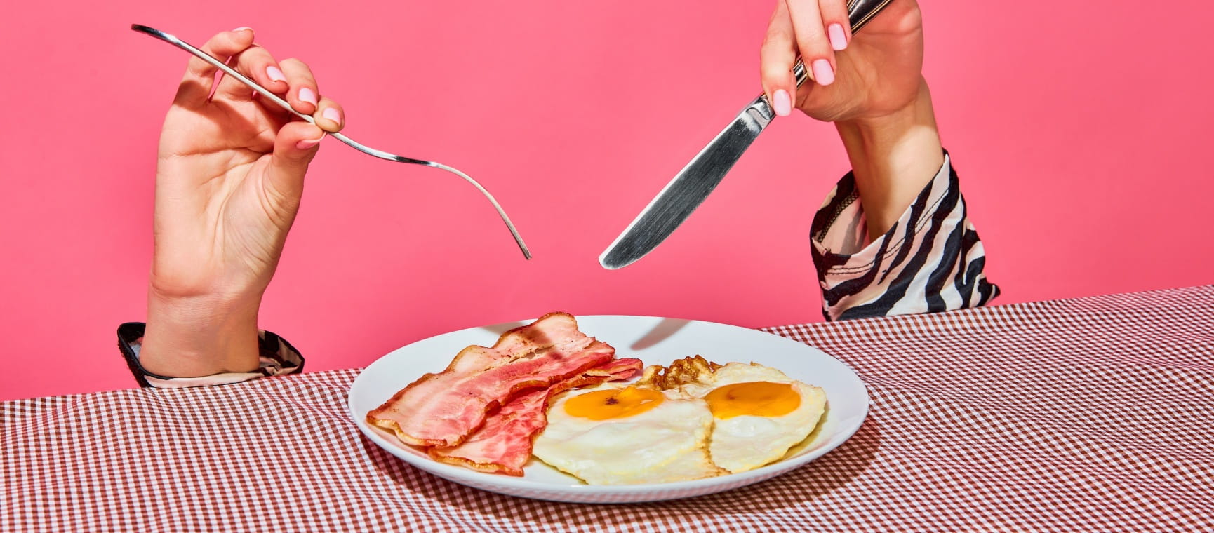 A pair of hands holding a knife and fork over a plate of eggs and bacon