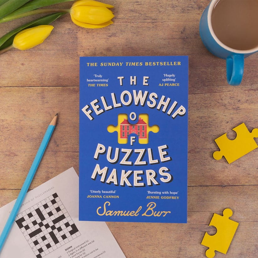 A copy of The Fellowship of Puzzle Makers on a wooden table next to puzzle pieces and a crossword