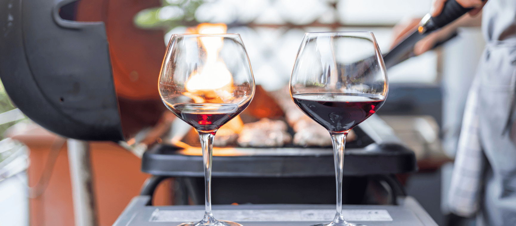 two glasses of red wine in front of a lit barbecue