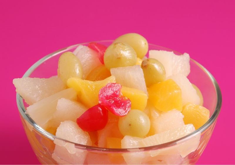 A clear bowl of tinned fruit cocktail against a bright pink background