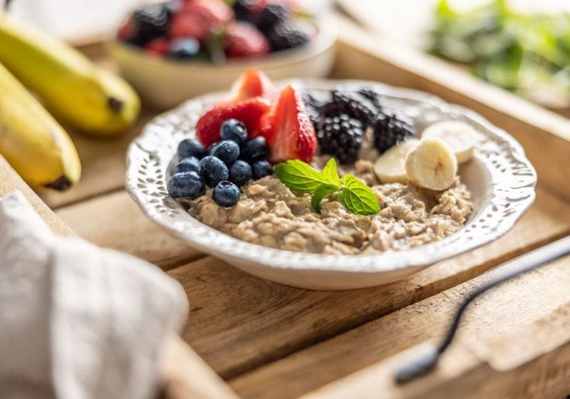 A bowl of porridge sits finished with berries and banana sits on a wooden tray