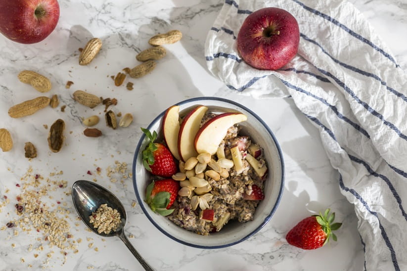 Bowl of oats porridge topped with sliced fresh fruit and nuts with strawberries and apple