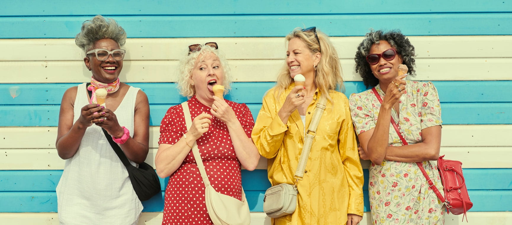 Four women laughing and leaning on a wall eating ice creams