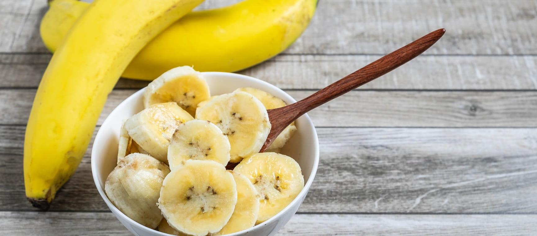 Unpeeled bananas on a wooden surface beside bananas cut up in a bowl