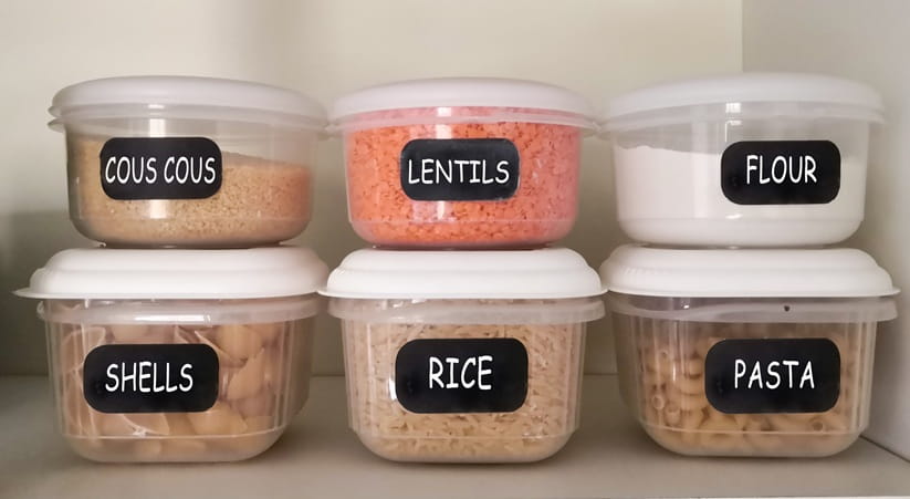 Plastic containers of dry food including couscous, red lentils, white flour, pasta, rice aligned on the kitchen cupboard shelf.