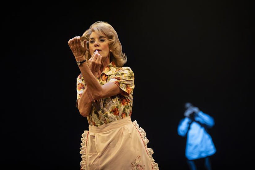 Tamzin Outhwaite in a floral collared dress with an apron putting on lipstick while holding up a compact mirror
