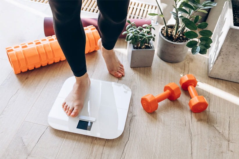 A person stepping on digital scales with a pair of orange dumbbells next to it on the floor