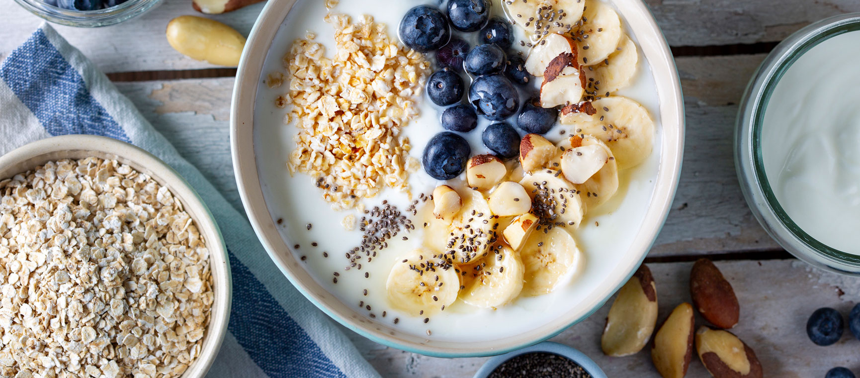 A bowl of porridge oats with bananas, blueberries, chia seeds and nuts on a table next to a bowl of dry oats and a pot of plain yoghurt