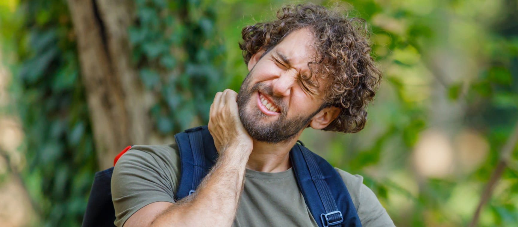 Man with beard in woods holding his neck in pain