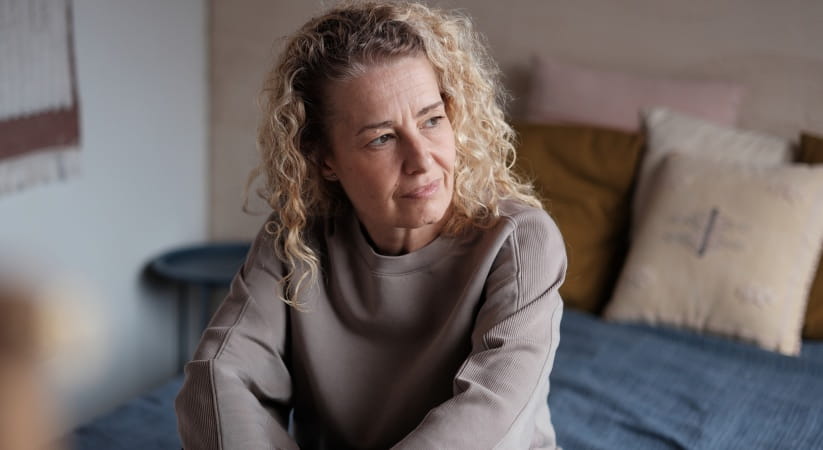Older woman with blonde curly hair sat on bed looking pensive