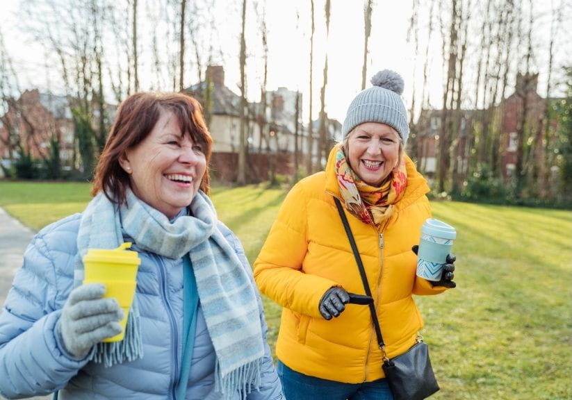 two women friends walking in the park in winter, laughing and carrying coffees