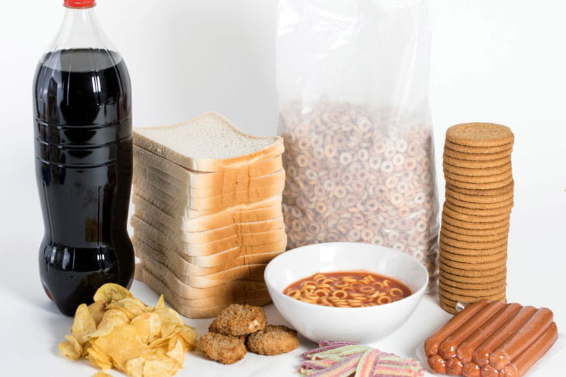 A selection of ultra processed food including white sliced bread, a bottle of soft drink, biscuits, frankfurters and biscuits