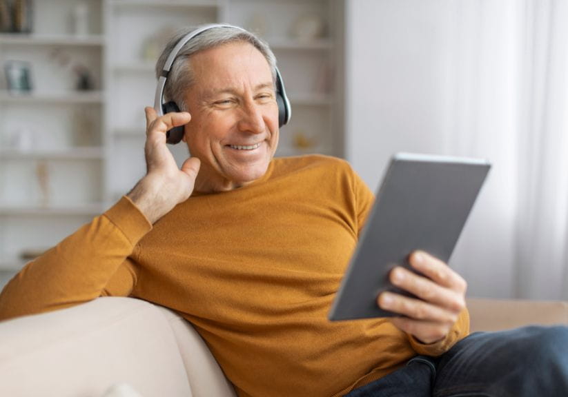 An older man sits on the sofa while listening to a podcast on a table through headphones
