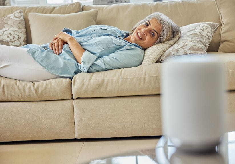 A mature lady lies on the sofa while listening to a Smart Speaker