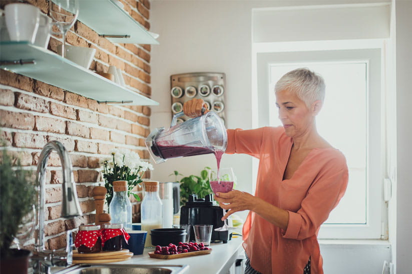 An older woman in a kitchen pouring a fruit smoothie into a glass