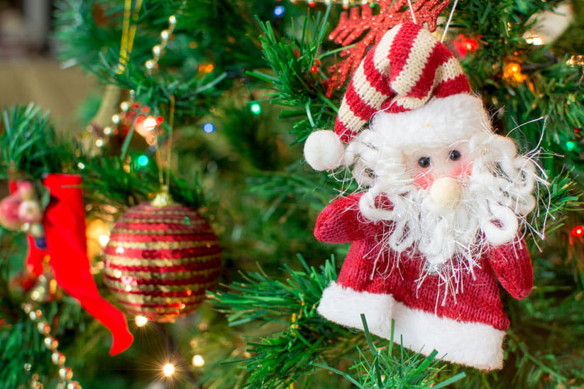 A hand-made, knitted Santa hanging from a Christmas tree branch with other decorations visible in the background