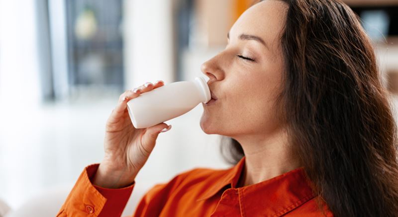 Side view of a woman in a rusty orange top drinking a bottle of probiotic yoghurt