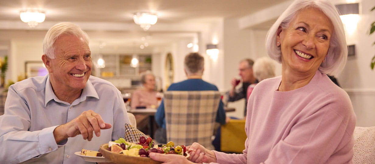 A smiling older man and woman sitting in a dining room sharing a food platter with people at another table behind them in the background