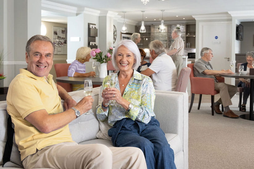 A group of older people in a communal dining hall enjoying glasses of bubbles