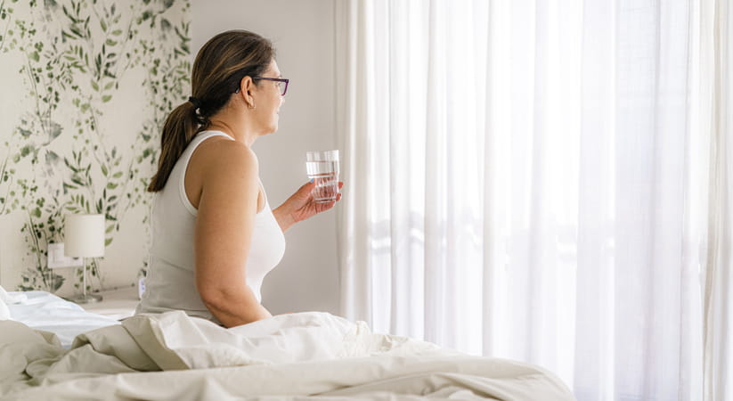 An older woman in white sitting on the edge of her bed drinking water
