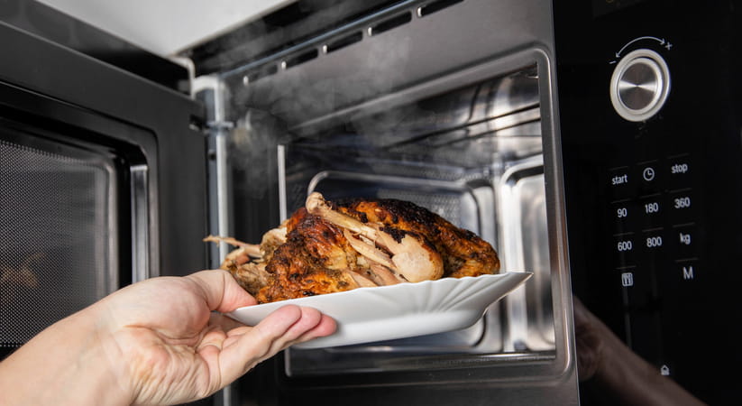 Close up of a hand putting a plate of cooked chicken into the microwave