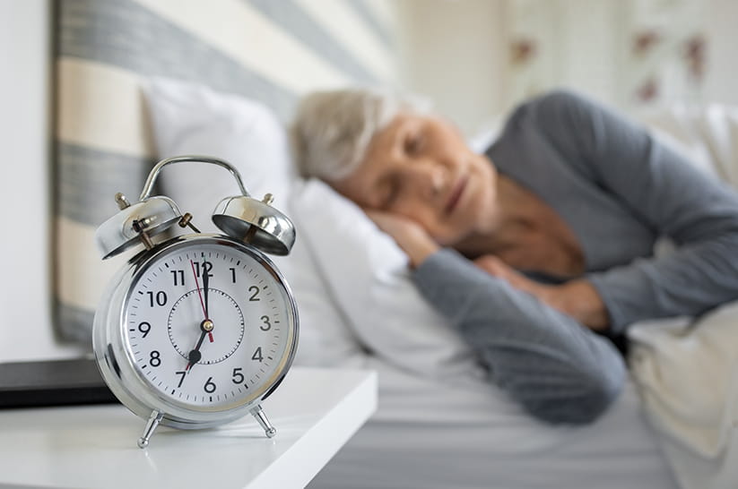 An older woman in bed with an alarm clock in the foreground