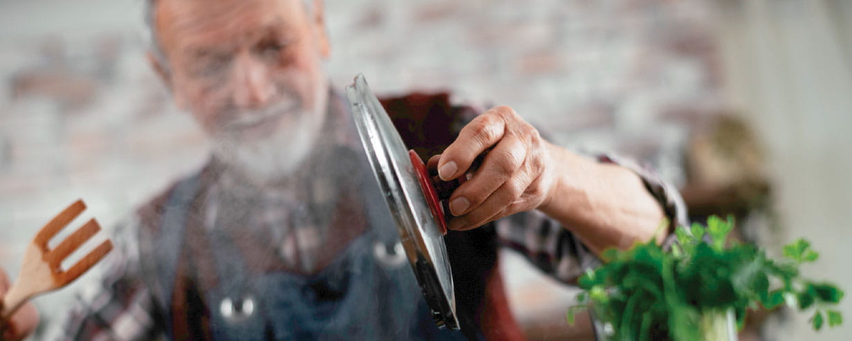Mature man in a kitchen, holding the lid of a pot with steam billowing up in front