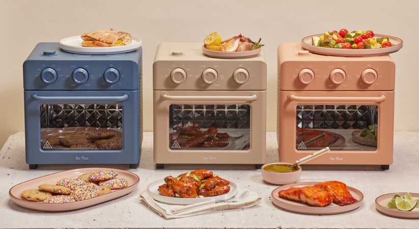Close up of three different coloured Our Place Wonder Ovens in a row with plates of food in front of them