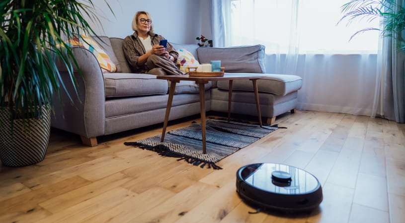 An older woman sits on the sofa operating a black robot vacuum cleaner with a remote control