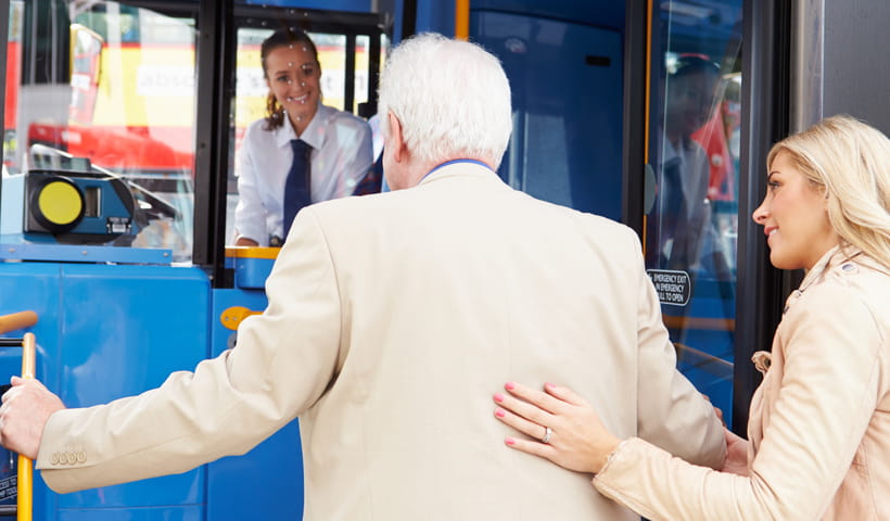Rear view of an older man in a cream jacket being helped onto a bus by a woman