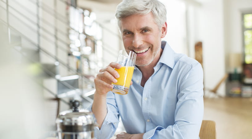 An older man in a light blue shirt smiles as he holds a glass of orange juice