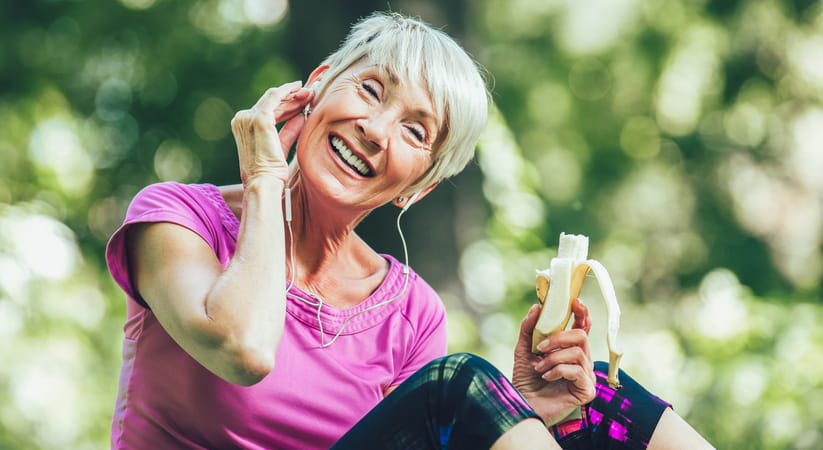 An older woman in a pink top smiles as she holds a half eaten banana