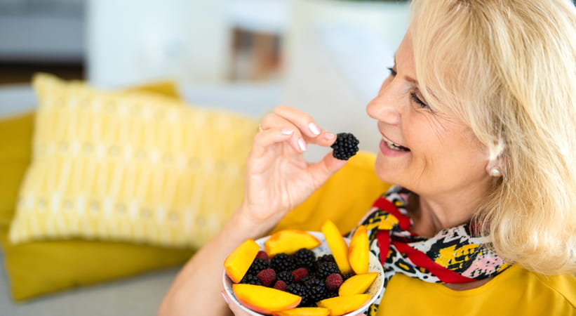 An older woman in a yellow top and a scarf smiles as she eats a bowl of fresh fruit