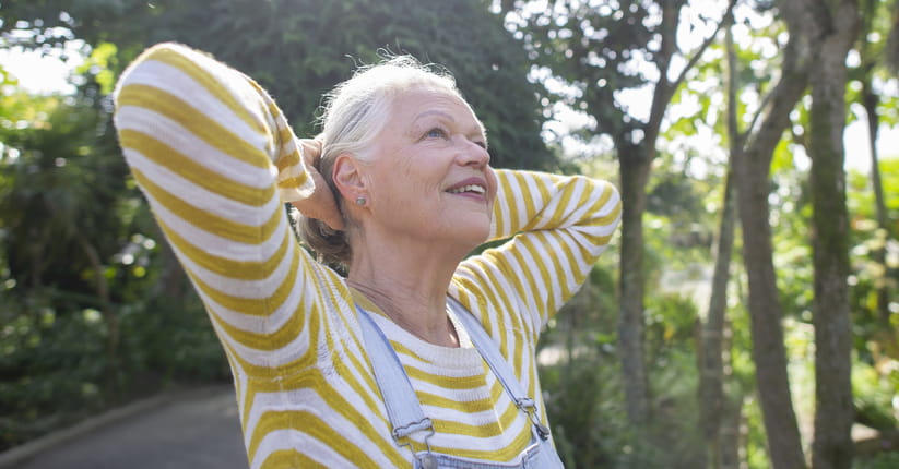 An older woman in a yellow hooped jumper and dungarees stands outdoors with her hands behind her head