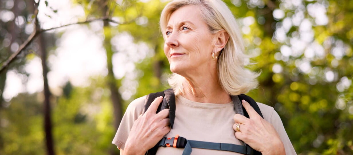 Close up shot of an older woman in a beige top and wearing a backpack standing in front of some trees