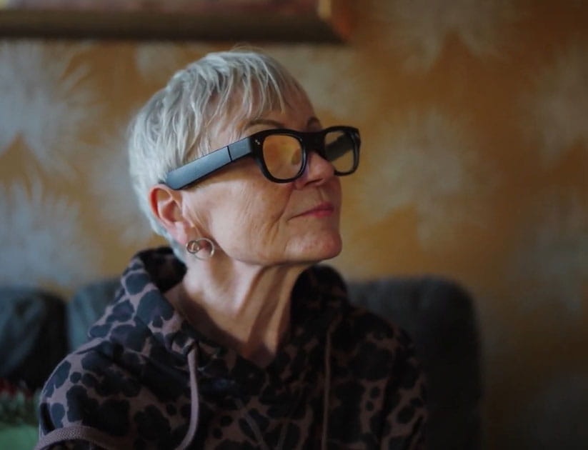 A profile view of a mature woman seated at home in her living room wearing the CrossSense demntia AI glasses