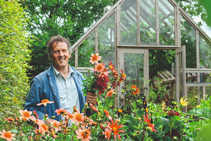 Monty Don standing in a garden, behind some flowers and in front of a greenhouse