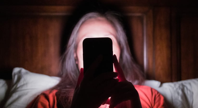 Woman looking at phone in bed. Her face is blocked by the phone and you can see the glow of the phone on her face
