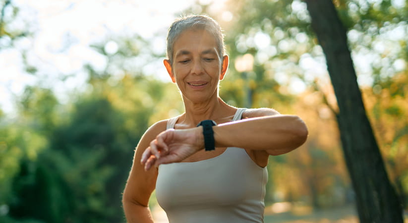 An older woman with short grey hair and a light coloured vest looks at her fitness band while exercising outdoors